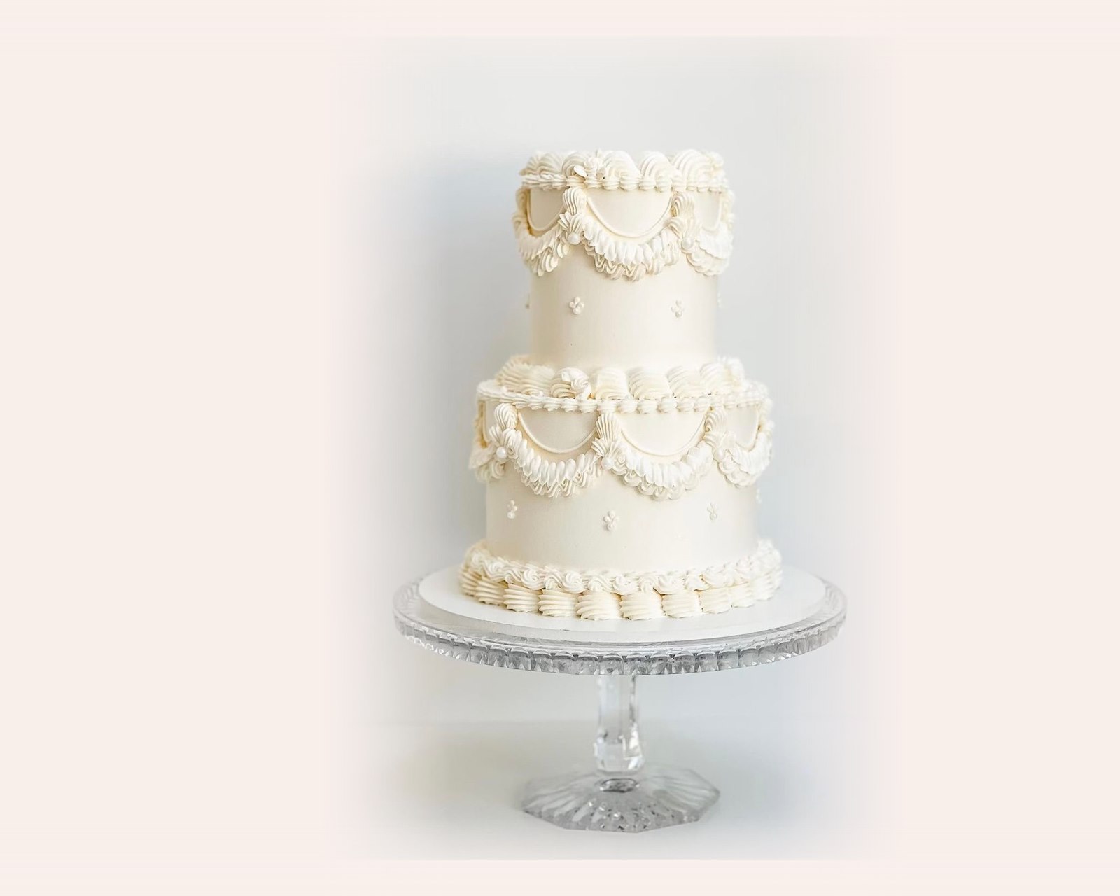 A white two tier celebration cake with piped Lambeth detail, on a glass cake stand